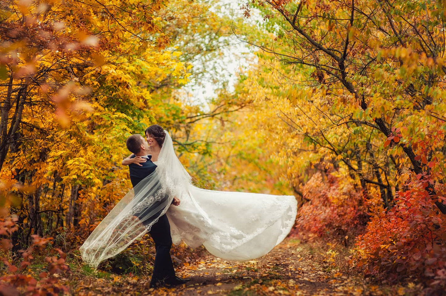 Newlywed couple. One lifitng the other up into the air. Standing in a forest path with vibrant fall leaves in the background.