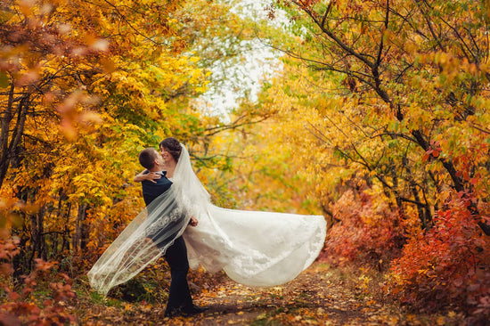 Newlywed couple. One lifitng the other up into the air. Standing in a forest path with vibrant fall leaves in the background.