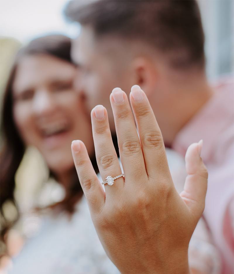 Close up photo of a hand wearing an engagement ring with a couple embracing in the background