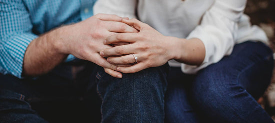 Photo of a couple sitting next to each other holding hands. One hand has a diamond engagement ring on it.